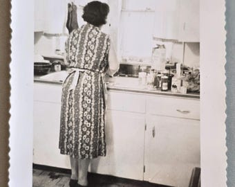 Foto vintage di una donna davanti al lavello della cucina, con calzini appesi sopra, vista posteriore e alimenti sul bancone.