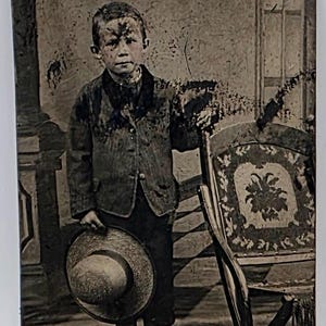 May include: A vintage black and white photograph of a young child in a jacket and hat, standing barefoot. The child is holding a straw hat and is standing next to a decorative chair. The image has a distressed appearance.