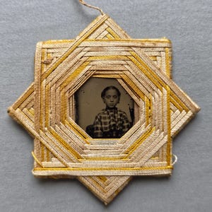 May include: A star-shaped frame constructed from woven beige and yellow thread, featuring a central octagonal opening that displays a vintage black and white photograph of a person wearing a plaid shirt. A thin string is attached to the top point.