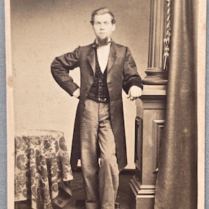 May include: A sepia-toned vintage photograph of a person standing in a formal frock coat, a patterned vest, and light-colored trousers. The person has a chin-strap beard and is positioned next to a draped curtain and a small table with a patterned cloth.