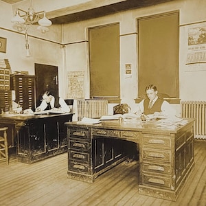 May include: Sepia-toned photograph of an office interior with two men seated at wooden desks, writing. The room features filing cabinets, framed pictures, and a vintage light fixture. A poster is visible on the wall. The scene evokes a sense of history and a bygone era.