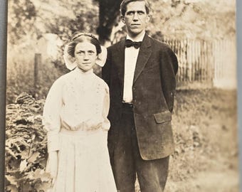 Chica RPPC con un gran lazo en el pelo y un hombre con pajarita, árbol y valla de madera al fondo.