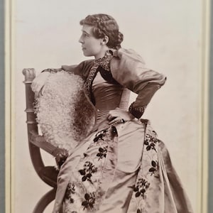 May include: A sepia-toned vintage photograph of a person in a high-collared dress with long sleeves and a floral patterned skirt, seated on a wooden chair. The bottom of the card features the handwritten text Cora Lingo Kelly and St. Louis.
