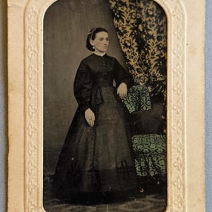 May include: A vintage black and white portrait of a woman in a long dress, seated near a patterned curtain and chair. The photograph is in a decorative frame, with handwritten text at the bottom. The woman is wearing a dark dress and has her hair styled up.