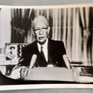 May include: Black and white photograph of a man in a suit and tie speaking at a podium. Two microphones are positioned in front of him. The background includes curtains and a glimpse of a framed picture.