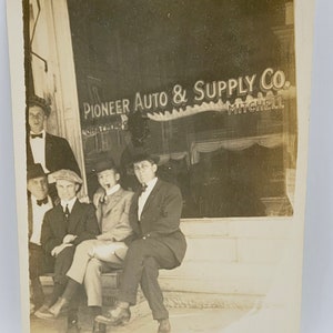 May include: Vintage black and white photograph featuring five men in suits and hats posing in front of a store with the sign "Pioneer Auto & Supply Co. Mitchell". The men are seated and standing, with a classic early 20th-century style.