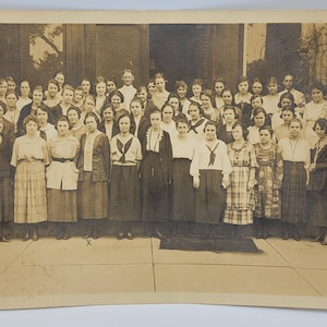 May include: A black and white vintage photograph of a group of women standing in front of a building. The women are all dressed in long dresses and have their hair pulled back. They are all smiling and looking at the camera.