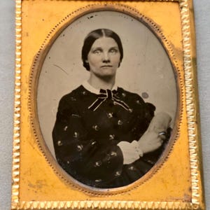 Puede incluir: Retrato ovalado enmarcado en un marco dorado. La persona del retrato lleva un vestido oscuro con cuello blanco y lazo. El marco tiene un borde decorativo. La persona mira directamente al espectador.