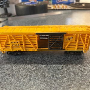 May include: Yellow model train boxcar with black lettering and a black door. The boxcar is labeled "M-K-T 47152" and "The Katy Serves the Southwest".