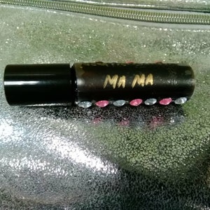 May include: A black rollerball perfume bottle with the gold text "MA MA". The bottle is decorated with pink and clear rhinestones. The bottle has a black cap and is set against a silver textured background.