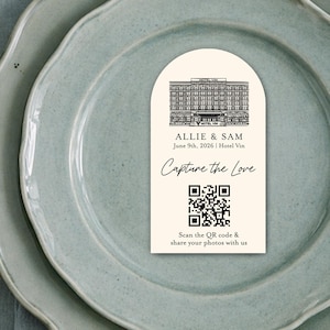 May include: A wedding table setting with a light blue plate, gold silverware, and a card that says "Capture the Love." The card has a QR code and a drawing of a building. Floral arrangement in the background.