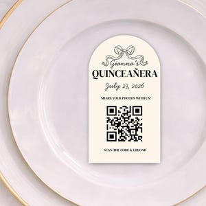 May include: A place setting with a white plate featuring a gold rim, gold silverware, and a cream-colored card. The card reads "Gianna's Quinceañera July 23, 2026" with a QR code and bow graphic.