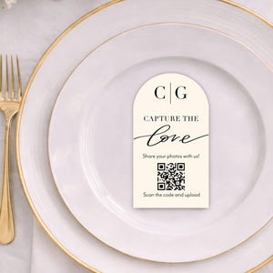 May include: A wedding place setting with a cream-colored card that reads "CAPTURE THE love" with a QR code. The card is on a white plate with a gold rim, next to gold silverware. Floral arrangement in the background.