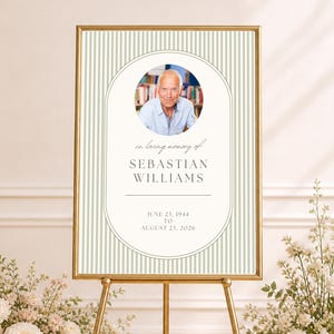 May include: A memorial sign on a gold easel with the text "in loving memory of Sebastian Williams". The sign features a photo and dates. Floral arrangements in pink and white flank the sign.