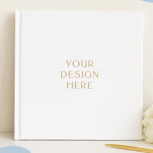 May include: A square white hardcover book with the gold foil text YOUR DESIGN HERE centered on the front cover. A gold pen lies horizontally on the surface next to a cluster of white peony flowers. The book features a clean, solid white finish.