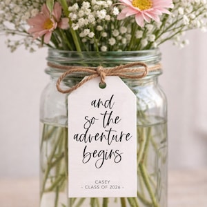 May include: A clear glass jar filled with water and stems, holding a bouquet of pink and white flowers. A white tag tied with twine reads "and so the adventure begins" with "CASEY - CLASS OF 2026 -" below.