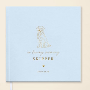 May include: A light blue memorial book with a gold illustration of a dog and the words "in loving memory" and "SKIPPER". The book also includes the years "2010-2026" and a paw print symbol.