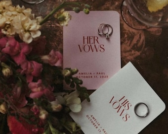 May include: Two pink cards with the text "Her Vows" and "His Vows" in red lettering. The cards are on a brown wooden surface with a bouquet of flowers and two glasses with lemon slices.