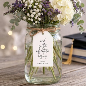 May include: A clear glass jar filled with water and fresh flowers, including white hydrangeas and purple statice. A white tag tied with twine reads "and so the adventure begins" with space for a name and "CLASS OF 2026".
