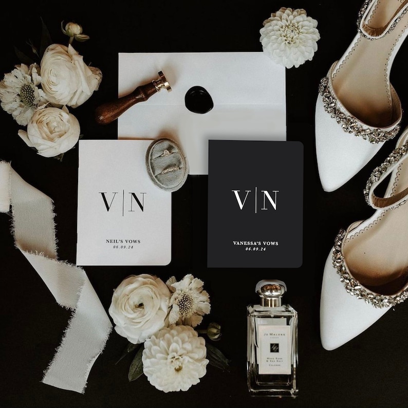 May include: A wedding flat lay on a black background. It features white bridal shoes, vow books with "VN" initials, a ring box, flowers, a perfume bottle, and a wax seal. The vow books are for Neil and Vanessa.