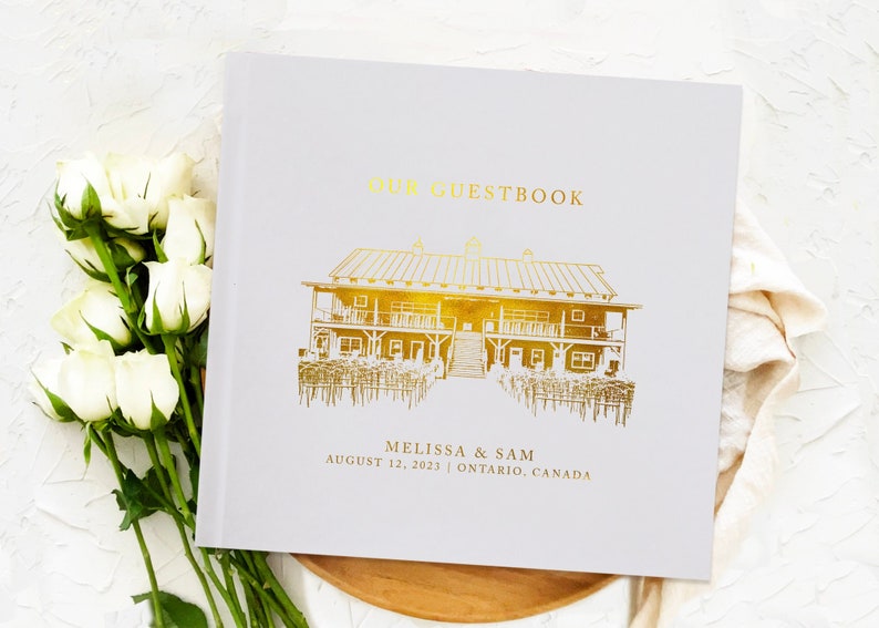 May include: A white guestbook with gold foil lettering and a gold illustration of a building. The text "OUR GUESTBOOK" is at the top, with names and a date below. A bouquet of white roses is on the left.