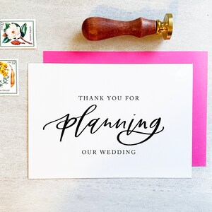 May include: A white card with black calligraphy that says "Thank you for planning our wedding". The card is on a pink cardstock and a wooden stamp with a gold top is in the upper right corner.