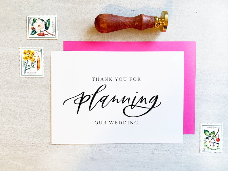 May include: A white card with black calligraphy that says "Thank you for planning our wedding". The card is on a pink cardstock and a wooden stamp with a gold top is in the upper right corner.