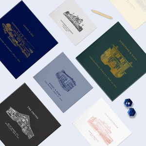 May include: An assortment of personalized guest books in various colors, including navy, white, and black. Each book features a custom illustration of a building and names with dates in gold, silver, or black ink. A wooden pen rests on top.