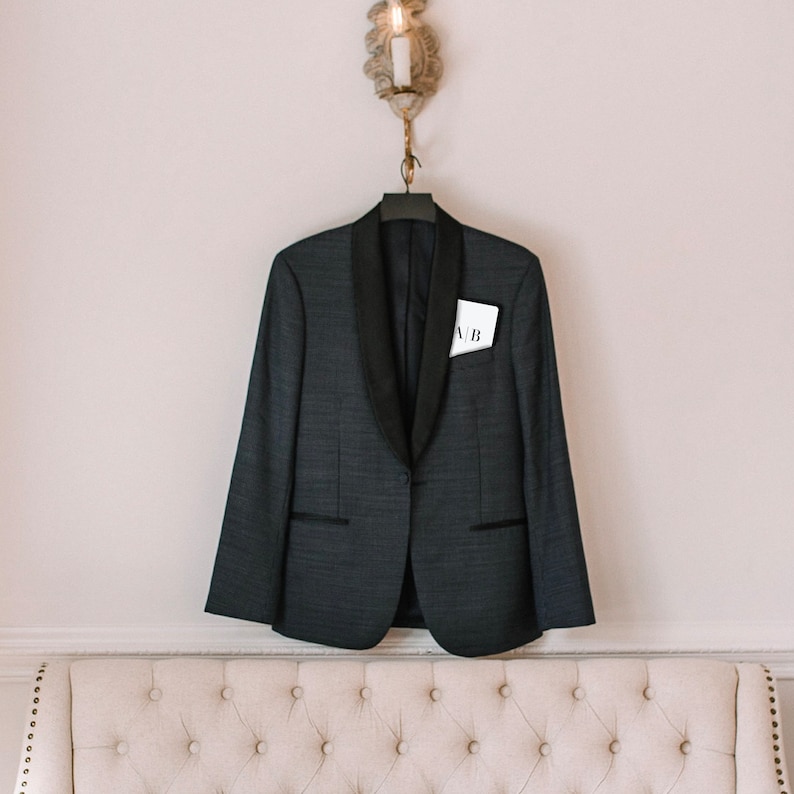 May include: A dark grey blazer with a black lapel and a pocket square displaying the initials "A/B" hangs on a hanger. The blazer is suspended on a wall, with a sconce above. This is a formal wear item.