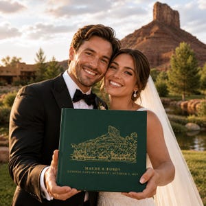 May include: A couple smiles, holding a dark green wedding album with gold foil details. The album features a landscape illustration and the names "Maude & Bobby" along with the wedding date and resort name. The background includes a mountain and water feature.