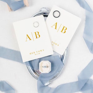 May include: A silver tray displays two cream vow cards with gold lettering, marked "A | B", "HER VOWS" and the date "09.9.20". Each card has a ring. A ring box and blue ribbon are also present, creating a wedding theme.