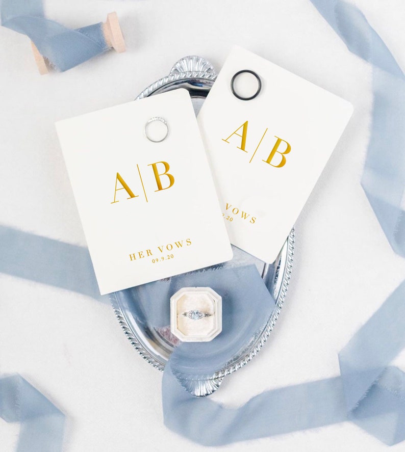 May include: A silver tray displays two cream vow cards with gold lettering, marked "A | B", "HER VOWS" and the date "09.9.20". Each card has a ring. A ring box and blue ribbon are also present, creating a wedding theme.