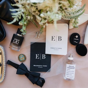 May include: A collection of wedding day items arranged on a surface. Includes a black bow tie, perfume bottles, vow books with "EB" initials, and a ring box with rings. A bouquet of white flowers is visible in the background. The vow books are for "Emily's Vows" and "Brannon's Vows".
