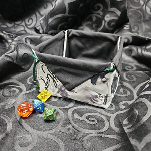 May include: A black and white fabric dice bag with green trim and the words "Good Luck" printed on the front. Four dice are scattered on a black fabric background with a swirling pattern.