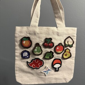 Peut inclure: Un sac fourre-tout en toile beige orné de onze écussons brodés. Les motifs incluent divers fruits, une feuille, une icône de fossile, un sac de clochettes, un nuage rouge, un symbole en éventail et une étoile à quatre branches sur le devant.