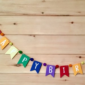 May include: A rainbow felt banner with the words "Happy Birthday" strung on a white cord with colorful pom-poms in between each letter.