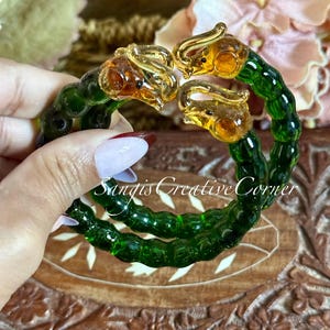 May include: A handcrafted glass bracelet featuring four dragon heads in amber and gold tones, connected by a green, beaded band. The bracelet is held against a wooden surface, with the text "Sangis Creative Corner" visible.