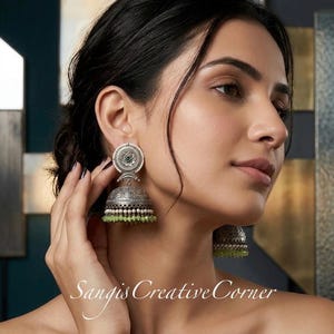 May include: Silver-toned jhumka earrings with a circular design and green bead accents. The earrings have a bell shape with intricate detailing and a small green stone in the center. The text "Sangis Creative Corner" is visible.