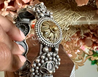 May include: A silver bangle bracelet with two decorative round medallions. The medallions have a raised design and are accented with gold. The bracelet is made of metal and has a textured surface.