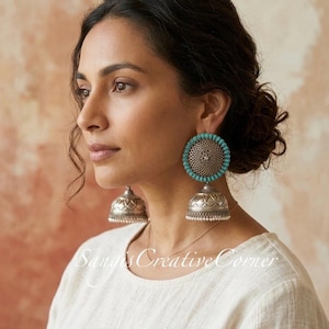 May include: Silver-toned jhumka earrings with turquoise accents and a detailed floral design. The earrings have a bell-shaped bottom and a circular top with turquoise beads. The earrings are worn by a person with dark hair and a white top.