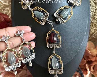 May include: A silver and gold necklace with a pendant featuring a red and white stone. The necklace has a geometric design and is accented with small pearl-like beads. Matching earrings with a similar design and stones are also included.
