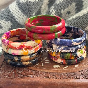 May include: A collection of colorful wooden bangles. The bangles are stacked in groups, showcasing a variety of patterns and colors, including red, green, blue, and floral designs. The text "Sam's Creative Corner" is visible.
