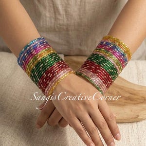 May include: A set of multi-colored glass bangles featuring a translucent finish with embedded gold-toned glitter accents. The bracelets are arranged in a stacked formation on each wrist, displaying a vibrant spectrum of red, green, blue, and yellow hues. The text Sangis Creative Corner is overlaid in white script.