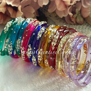 May include: A collection of colorful bangles in shades of green, teal, pink, purple, blue, yellow, and red. Each bangle is adorned with sparkling rhinestones. The bangles are arranged in a row, showcasing their vibrant colors and intricate details.