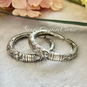 May include: Two silver bangles with intricate floral designs. The bangles are stacked on top of each other and have a shiny finish.