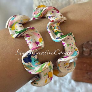 May include: Two abstract, cream-colored bracelets with a colorful splatter paint design. The bracelets are in a wavy, curved shape, with pink, yellow, blue, and green paint splatters. The text "San's Creative Corner" is visible.