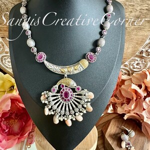 May include: A silver and gold necklace with a pink stone pendant. The pendant is shaped like a fan and has a fish design. The necklace is on a black stand and there are matching earrings in the background.