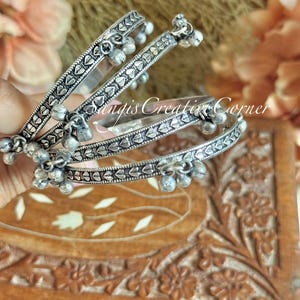 May include: Four silver bangles with intricate heart and leaf patterns and small dangling bells. The bangles are displayed on a wooden surface with floral carvings, creating a rustic and elegant aesthetic.