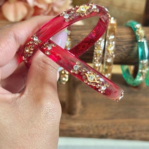 May include: A pair of red bangles with gold and crystal embellishments. The transparent red bracelets are adorned with gold-toned metal and clear crystals. Other bangles in gold and green are visible in the background, displayed on a wooden stand.