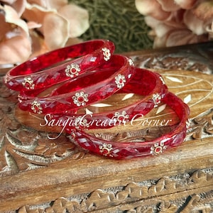 May include: A stack of five faceted red bangles with floral embellishments. Each bangle features a repeating pattern of small, gold-toned flowers with red center stones. The bangles are displayed on a carved wooden surface.